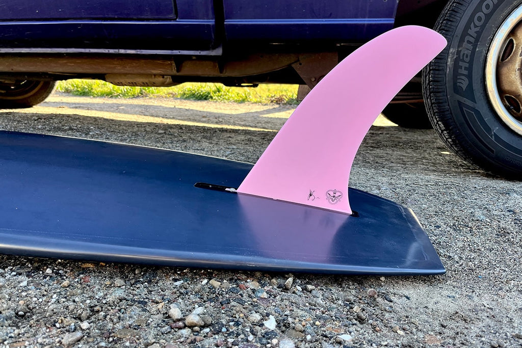 Pink Phase 3 fin on a navy longboard in the parking lot before a surf.